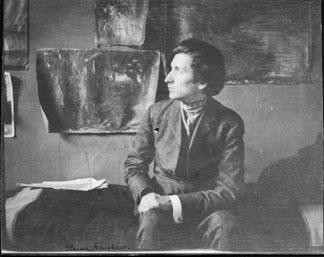 Photograph of Glen Cooper Henshaw seated in his studio, with canvases pinned to the wall behind him